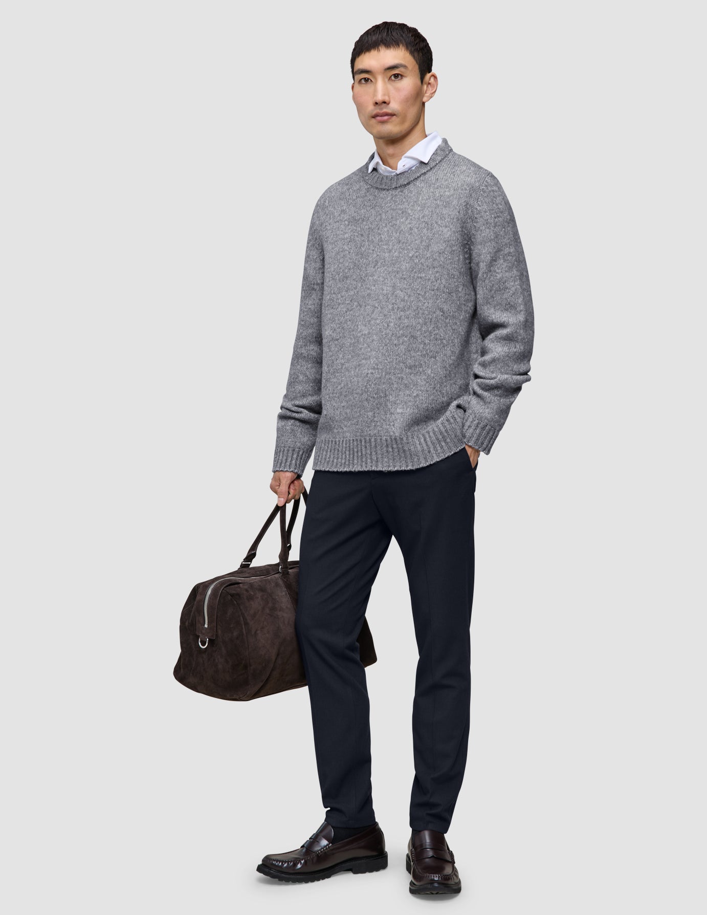 En mand iført en grå sweater, en hvid skjorte og et par Midnight Blue Motion-bukser i slim-fit, der holder en brun rejsetaske.